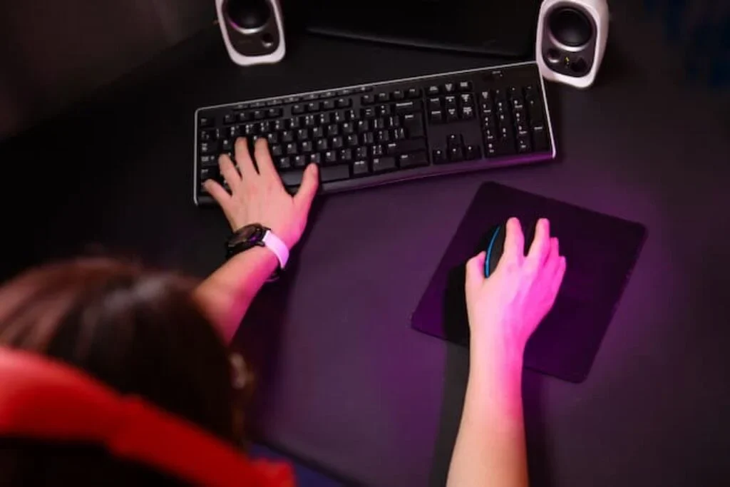 UMA PESSOA JOGANDO COM AS MÃOS UMA NO TECLADO E OUTRA NO MOUSE E AO FUNDO DUAS CAIXAS DE SOM BRANCA.